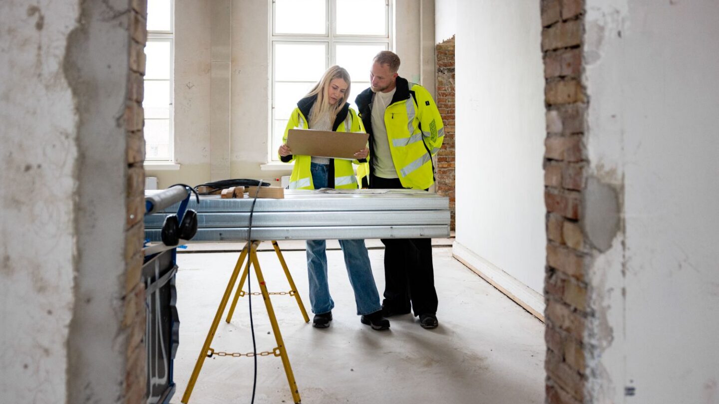 To bygningskonstruktører iført gule veste. De kigger på bygningstegninger.